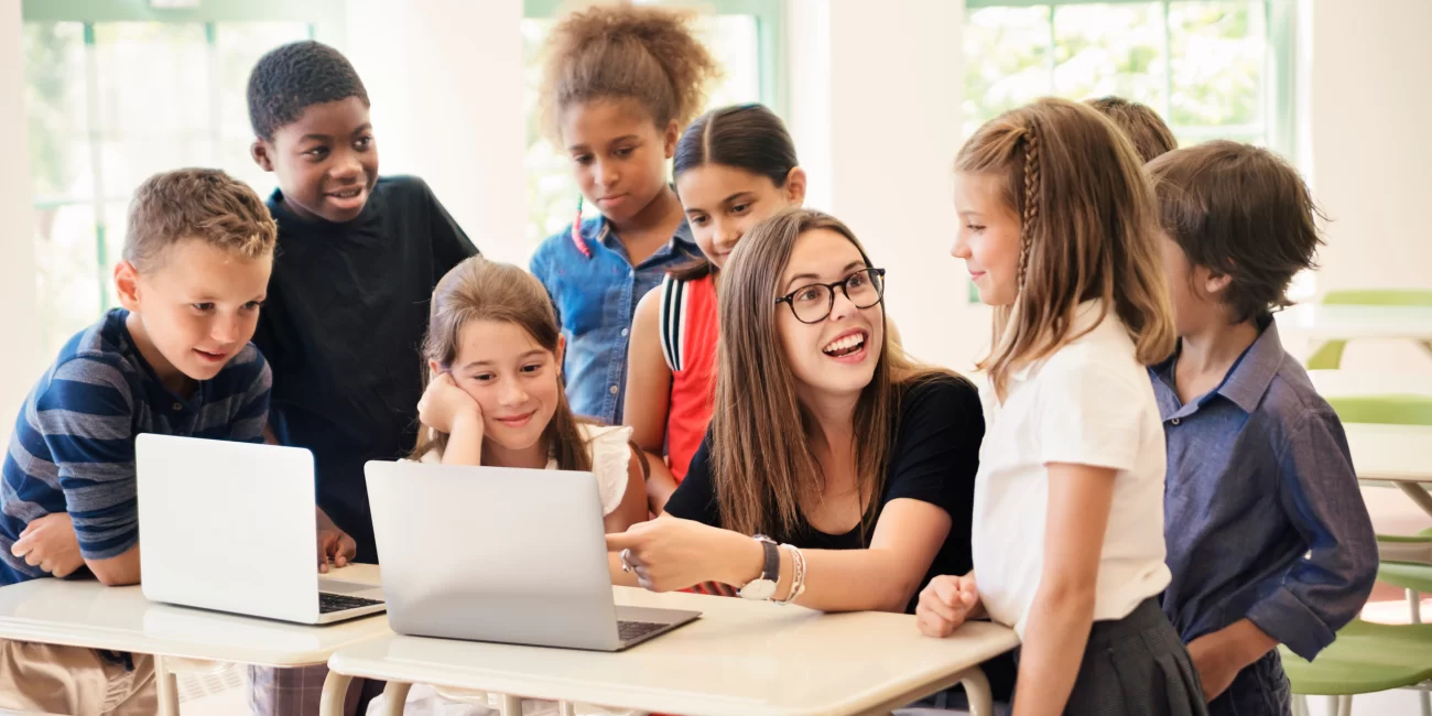Learning Lands – Our award-winning online learning platform: this is a photo of a teacher surrounded by students, showing them something on her laptop